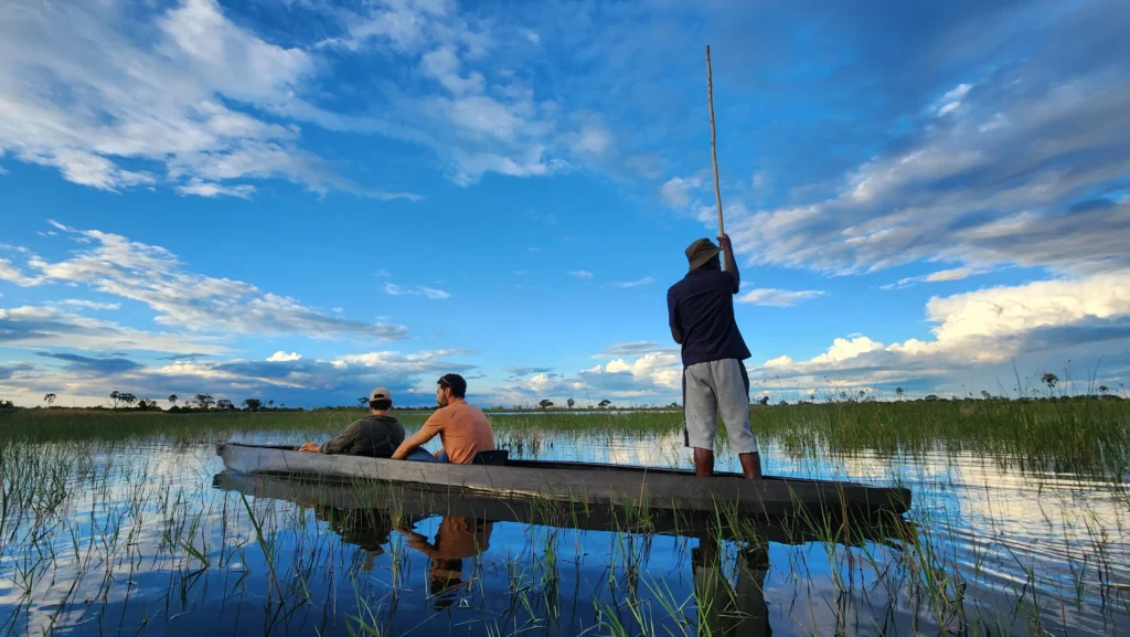 Two tourists sit in a traditional mokoro dugout canoe while a guide stands and poles through the calm waters of the Okavango Delta under a vibrant blue sky.