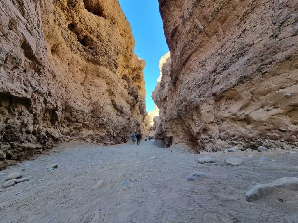 A low-angle view looking through the narrow, towering sandy-colored rock walls of Sesriem Canyon in Namibia, with a few tourists walking in the distance.