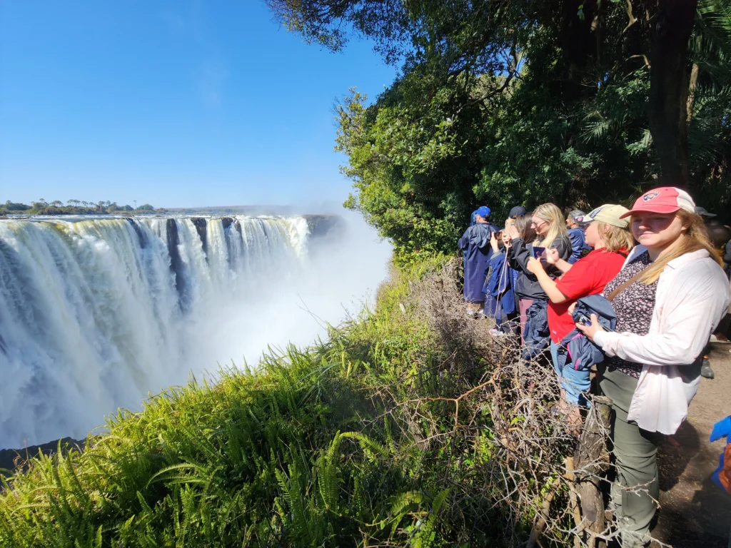 A group of tourists standing on a lush, green viewing platform taking photos of the massive, cascading Victoria Falls under a clear blue sky.